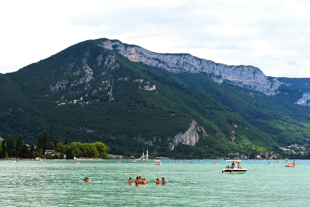 Vue sur le lac d’Annecy en été avec baigneurs et pédalos, entouré des montagnes verdoyantes